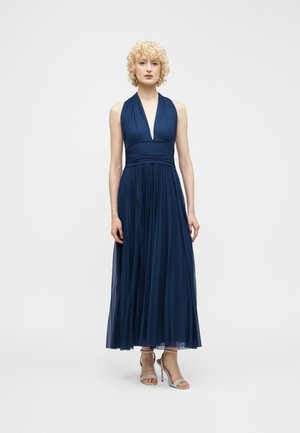 Woman with curly blonde hair wearing a sleeveless navy blue pleated midi dress and beige high-heeled sandals, standing against a plain background.