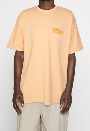 T-shirt à manches courtes de couleur pêche en coton, avec un graphic imprimé jaune qui indique "On Vacation Club" sur le côté gauche de la poitrine.