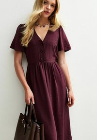 Robe bordeaux à mi-longueur avec des manches courtes, détail de boutons, tissu texturé et taille cintrée. Associée à un sac à main marron.
