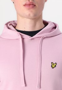 Hoodie rose en tissu doux avec une poche frontale. Dispose d'une capuche avec cordon de serrage et d'un petit logo jaune brodé sur la poitrine.