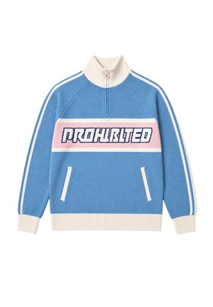 Mand iført en blå og hvidstribet sweater med teksten "Honrlee", mørke bukser og en grå strikket hue, står mod en ensfarvet baggrund.