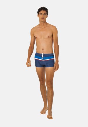 Jeune homme marchant pieds nus portant un short de bain bleu avec des rayures blanches, bleu clair et rouges sur un fond uni.
