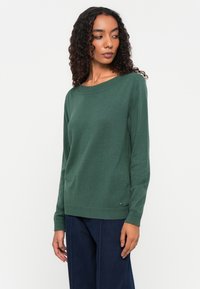 Green knitted sweater with long sleeves, round neckline, and ribbed hem. Features a small logo detail at the bottom left side.