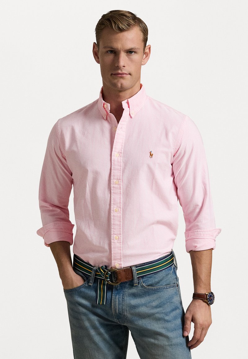 Camisa de botão rosa às riscas, com mangas longas, fabricada em material suave e com um pequeno logótipo como detalhe. Usada com jeans azuis e um cinto multicolorido.