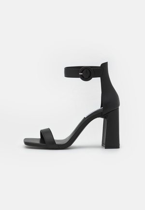 Czarne sandały na wysokim obcasie z zamkniętym tyłem, paskiem na kostkę z okrągłą klamrą i szerokim słupkowym obcasem. Wykonane z gładkiej skóry, o minimalistycznym designie.