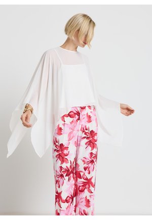 Frau mit kurzen blonden Haaren, die ein lockeres, weißes, durchsichtiges Oberteil und weit geschnittene Hosen mit großem rosa und rotem Blumenmuster trägt.