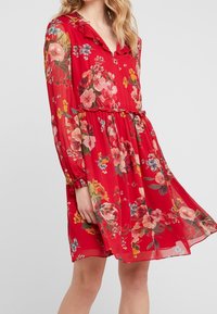 Robe fleurie rouge à manches longues, avec un col froncé et une taille cintrée. Présente des fleurs vibrantes en rose, jaune et bleu sur un tissu transparent.