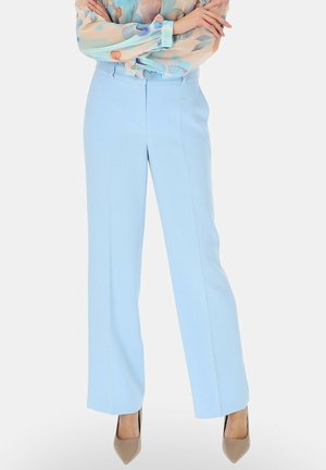 Osoba ubrana w jasnoniebieskie, dopasowane spodnie z wysokim stanem, beżowe czółenka na obcasie oraz pastelową, kwiecistą bluzkę z długim rękawem, z założonymi ramionami.