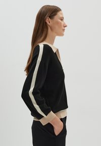 Maglione nero con scollo a V, colletto beige e strisce bianche sporche lungo le maniche. Texture morbida e orlo a coste. Indossato con pantaloni neri.