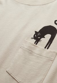 Camiseta de algodón beige claro con un gráfico negro de un gato en la parte izquierda del pecho, con un bolsillo de acabado texturizado.