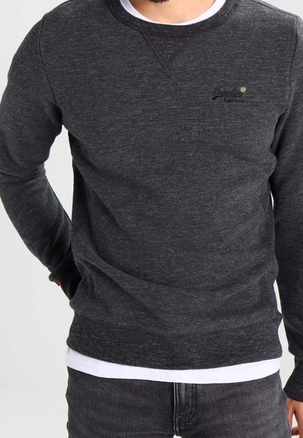 Homme portant un sweat-shirt gris foncé à manches longues et col rond par-dessus une chemise blanche, avec un jean gris foncé, debout avec un bras derrière le dos.
