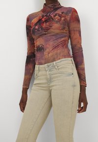 Haut à manches longues avec un motif artistique coloré en violet, orange et jaune ; doté d'un col montant et d'un détail froncé sur le côté. Associé à un jean en denim clair.