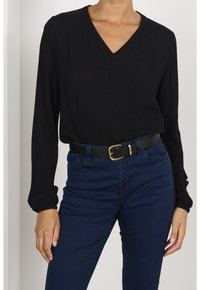 Femme portant un blouse noire à manches longues et décolleté en V, rentrée dans un jean bleu foncé avec une ceinture en cuir noire ornée d'une boucle dorée.