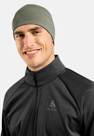 Olivgrüner Beanie aus dehnendem Stoff, eng anliegendes Design, glatte Textur. Getragen mit einer schwarzen Zip-Jacke mit einem Logo auf der Brust.