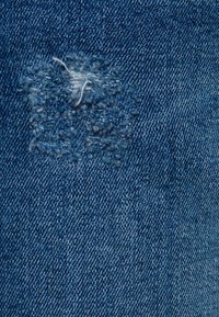 Denimová látka s centrálními, nepravidelnými nášivkami ve světle modré barvě, s viditelnou strukturovanou texturou a volnými nitěmi.