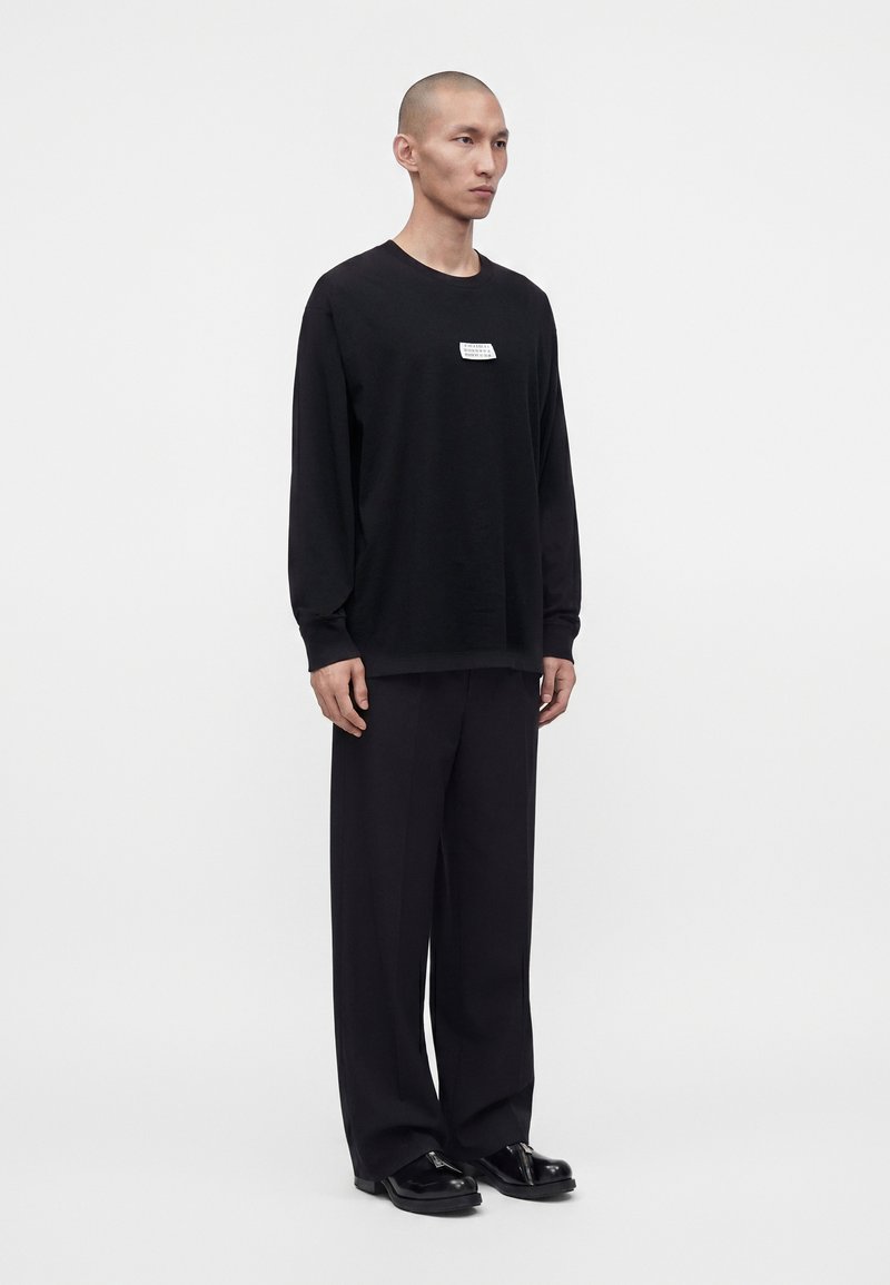 Mann mit rasiertem Kopf steht frontal, trägt ein schwarzes Langarmshirt mit weißem Brustlogo, schwarze weite Hose und schwarze Schuhe vor einfachem Hintergrund.