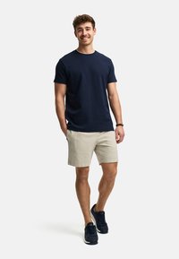 Camiseta de algodón navy, pantalones cortos beige texturizados y zapatillas oscuras. El modelo se encuentra de pie de manera casual con las manos en los bolsillos, sonriendo frente a un fondo blanco.