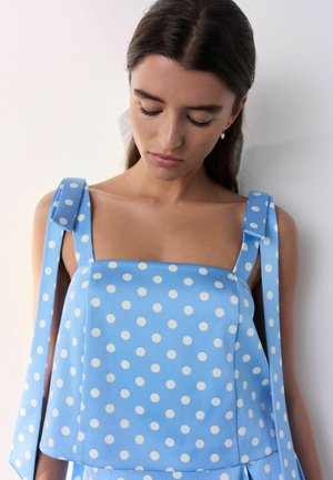 Fiatal, sötét hajú nő, kék, ujjatlan pöttyös felsőt visel, széles vállpántokkal, amelyeket masnik kötnek össze, lefelé néz.