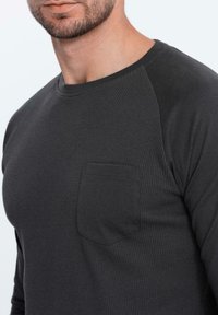 Schwarzes Langarmhemd mit gerippter Textur, das eine Brusttasche hat. Der Stoff hat eine glatte Verarbeitung an den Ärmeln und dem Kragen.