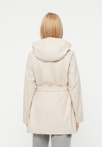Imperméable beige clair avec capuche, taille ceinturée et manches longues. Texture lisse et design simple, se terminant au milieu des cuisses.