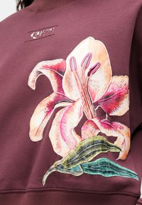 Sudadera color marrón con una gran flor de lirio bordada en rosa y blanco, y hojas verdes. Pequeño logo de Guess encima de la flor.