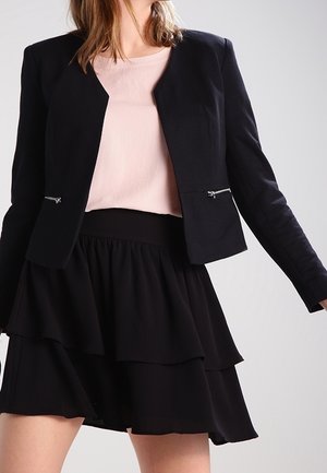 Frau trägt einen schwarzen Blazer mit Reißverschlusstaschen, ein hellrosa Top und einen geschichteten schwarzen Rock vor einfachem hellem Hintergrund.