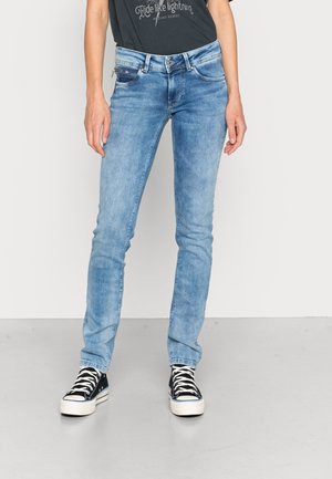 Person, die hellblaue Slim-Fit-Jeans und schwarze High-Top-Sneaker mit weißen Schnürsenkeln trägt, steht vor einem schlichten hellen Hintergrund.