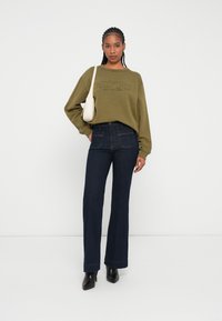 Felpa verde oliva con logo ricamato, abbinata a pantaloni jeans a gamba larga blu scuro. Il modello tiene una piccola borsa bianca e indossa tacchi neri.