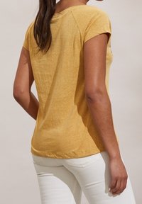 Mujer con cabello largo y oscuro, vestida con una camiseta amarilla de manga corta y pantalones blancos, vista desde atrás contra un fondo sencillo.