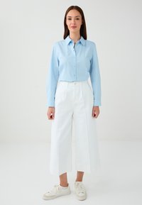Woman standing wearing a light blue button-up shirt, white wide-leg cropped pants, and white sneakers against a plain white background.