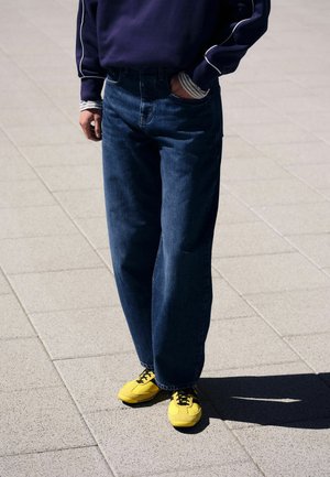 Person standing on a tiled pavement wearing dark blue jeans, a navy sweater with white trim, and bright yellow sneakers with black laces.
