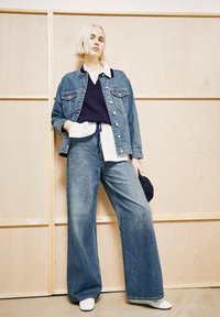 Woman with short blonde hair wearing layered denim jacket and wide-leg jeans, holding a navy cap, standing against wooden panel background.