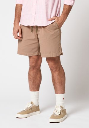 Man wearing beige drawstring shorts, white socks, beige sneakers, and a pink button-up shirt with sleeves rolled up.