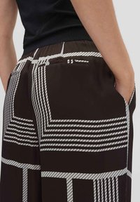 Bruine broek met een geometrisch wit patroon. Inclusief zijzakken en een gladde, lichte stof met een elastische tailleband.