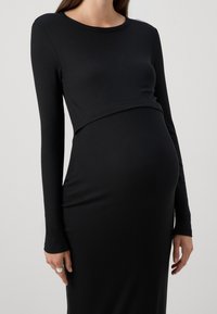 Svart långärmad gravidklänning i ribbad design, figurnära passform som accentuerar en rundad mage, slät textur och subtil sömdetaljering.
