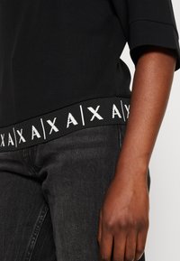 Černé tričko s krátkým rukávem, které má žebrovaný pas s bílým geometrickým logem. V kombinaci s tmavě šedými kalhotami, záběr na ruku.