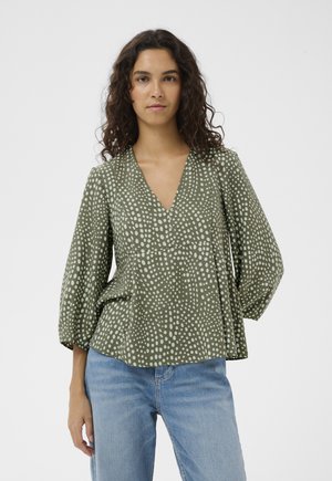 Femme aux cheveux bouclés portant un chemisier à manches longues à pois vert olive et un jean bleu clair, debout devant un fond blanc.