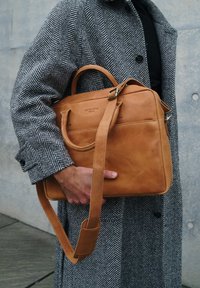 Tan leather bag with a structured design, detachable shoulder strap, and top handles. Features a front pocket and embossed logo.