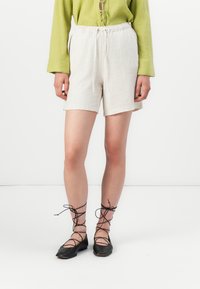Beige linnen shorts met een elastische tailleband en een koord. Gecombineerd met zwarte veterschoenen en een structuur groen shirt.