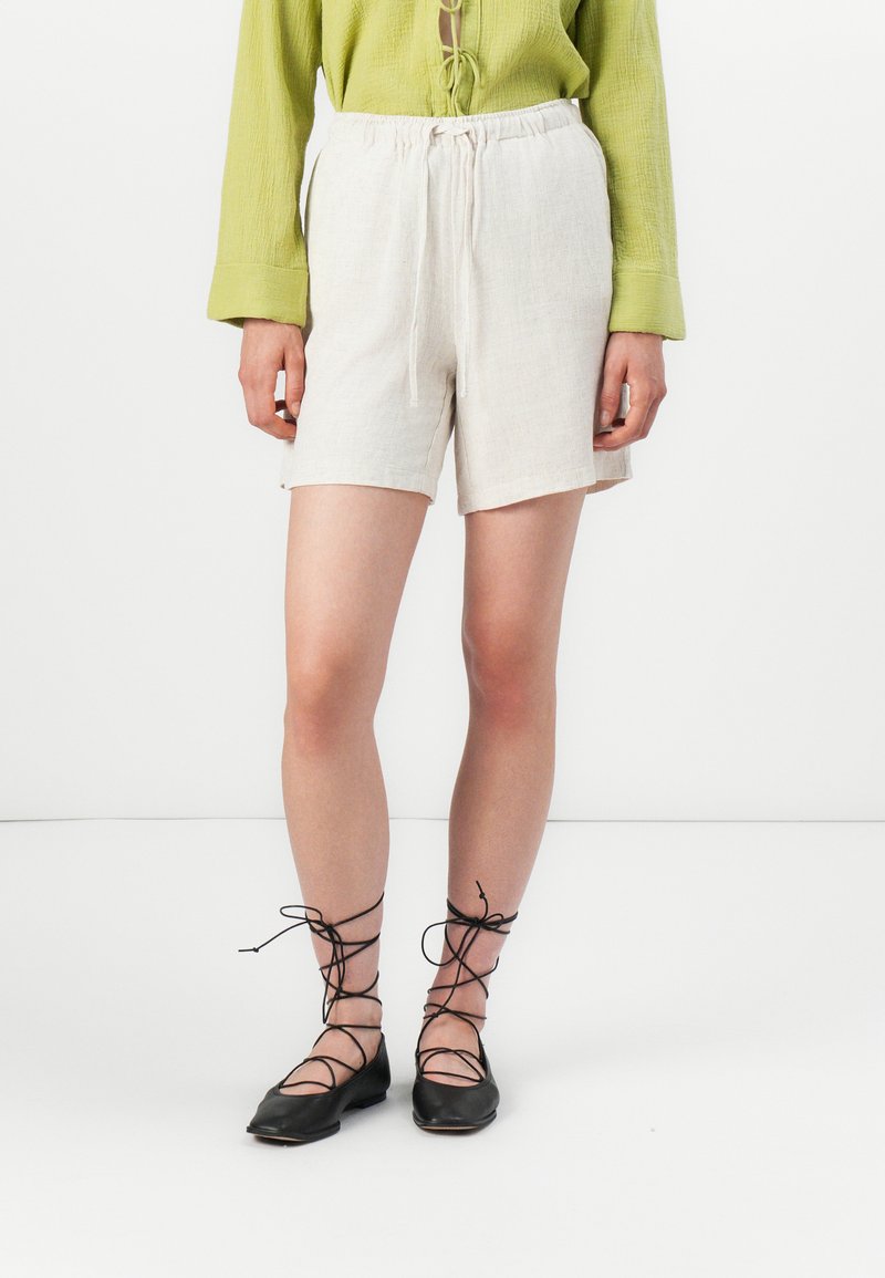 Beige linnen shorts met een elastische tailleband en een koord. Gecombineerd met zwarte veterschoenen en een structuur groen shirt.