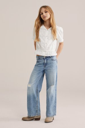 Jauna mergina su ilgais šviesiais plaukais, vilkinti balta tekstūruota palaidine, platėjančiais mėlynais suplyšusiais džinsais ir smėlio spalvos kulkšnies ilgio batais, stovinti su rankomis už nugaros.