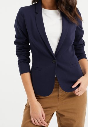 Femme portant un blazer bleu marine cintré sur un top blanc et un pantalon marron clair, debout avec une main dans la poche et l'autre posée sur la hanche.
