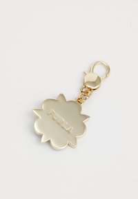 Porte-clés en forme de fleur dorée avec un fermoir, gravé avec "FURLA" au centre, sur un fond blanc uni.