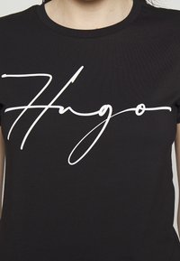 Schwarzes Baumwoll-T-Shirt mit großem, weißem, geschwungenem Logo "Hugo", das prominent auf der Brust gedruckt ist. Kurze Ärmel, runder Ausschnitt.