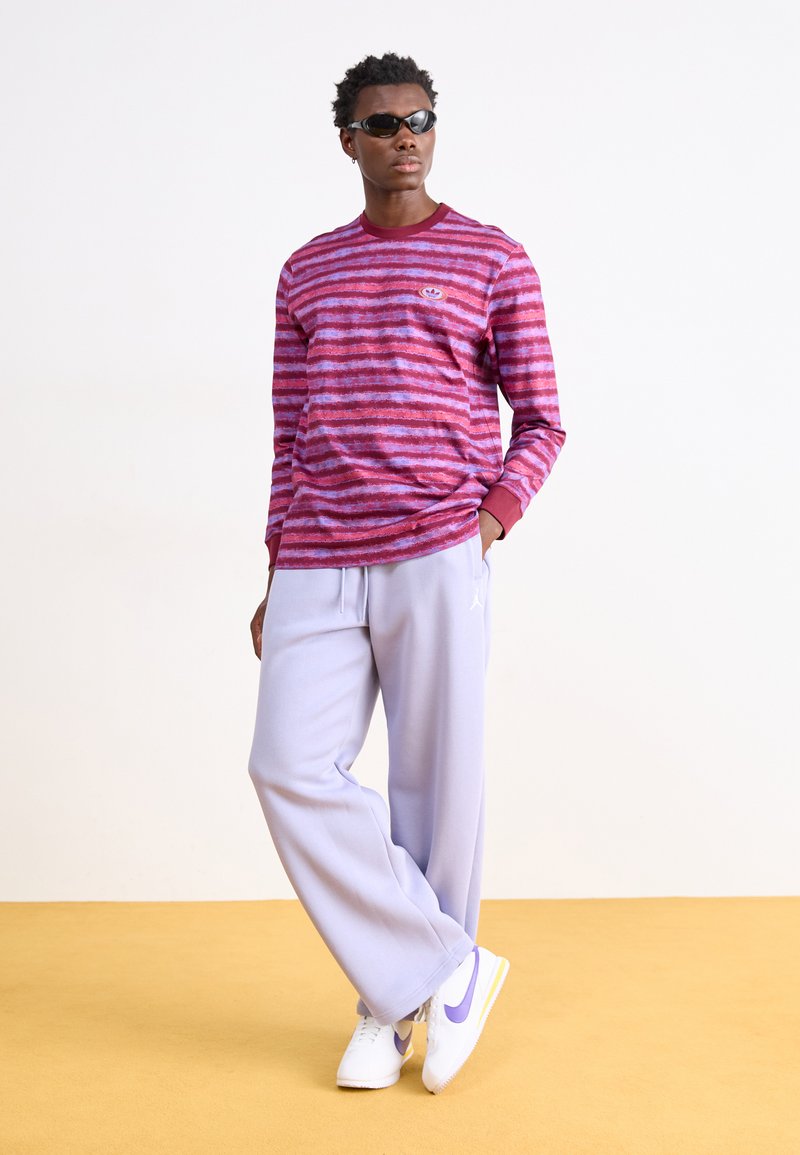 Chemise rayée bordeaux et violette à manches longues avec col rond, associée à un pantalon de survêtement violet clair ample et des baskets blanches avec des accents violets.