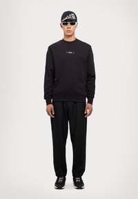 Felpa nera con un patch del logo, abbinata a pantaloni leggeri neri. Gli accessori includono un cappellino nero e occhiali da sole bianchi. Le sneakers sono nere con dettagli bianchi.