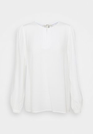 Blouse blanche à manches longues en tissu subtilement texturé, col rond et manches légèrement bouffantes terminées par des poignets.