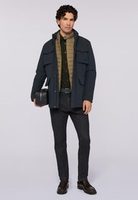 Giacca esterna blu navy, strato interno imbottito beige, camicia verde, pantaloni in denim scuri, cintura nera e mocassini marroni; presenta numerose tasche.