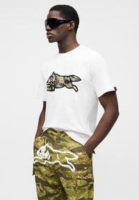 Λευκό μπλουζάκι με μαύρο και καμουφλάζ γραφικό σχέδιο· πράσινο παντελόνι καμουφλάζ cargo με λευκή φιγούρα δρομέα. Μαύρα γυαλιά ηλίου.