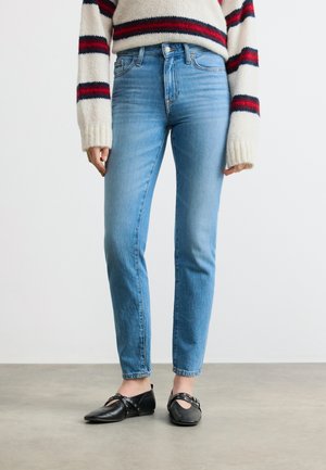 Žena stojaca na sivej podlahe, oblečená v svetlomodrých úzkych džínsoch, krémovom svetri s červenými a čiernymi pruhmi a čiernych špicatých balerínach so pásikmi.